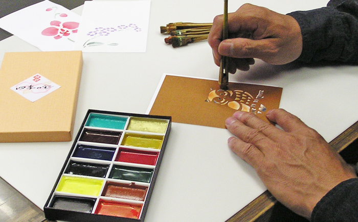 手作り年賀状を型紙を使って作成（顔彩絵の具と刷毛で彩色）している場面
